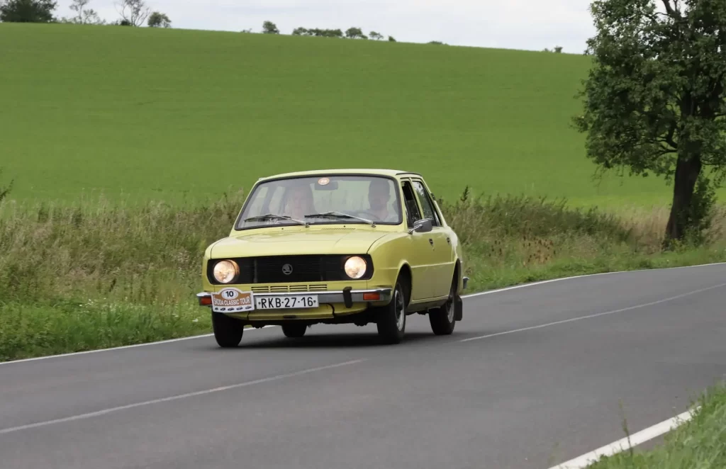 soc3 - Techsvět Starší žlutá Škoda 105, která projíždí klikatou silnicí mezi poli.
