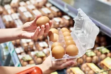 Žena v obchodě drží otevřenou plastovou krabičku s deseti hnědými vejci.