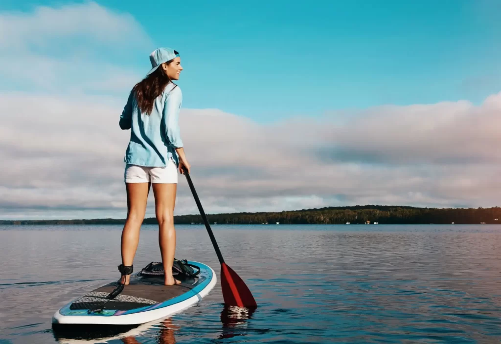 vlny1 - Techsvět Mladá žena v modré košili a bílých šortkách stojí na paddleboardu na klidné vodní hladině.