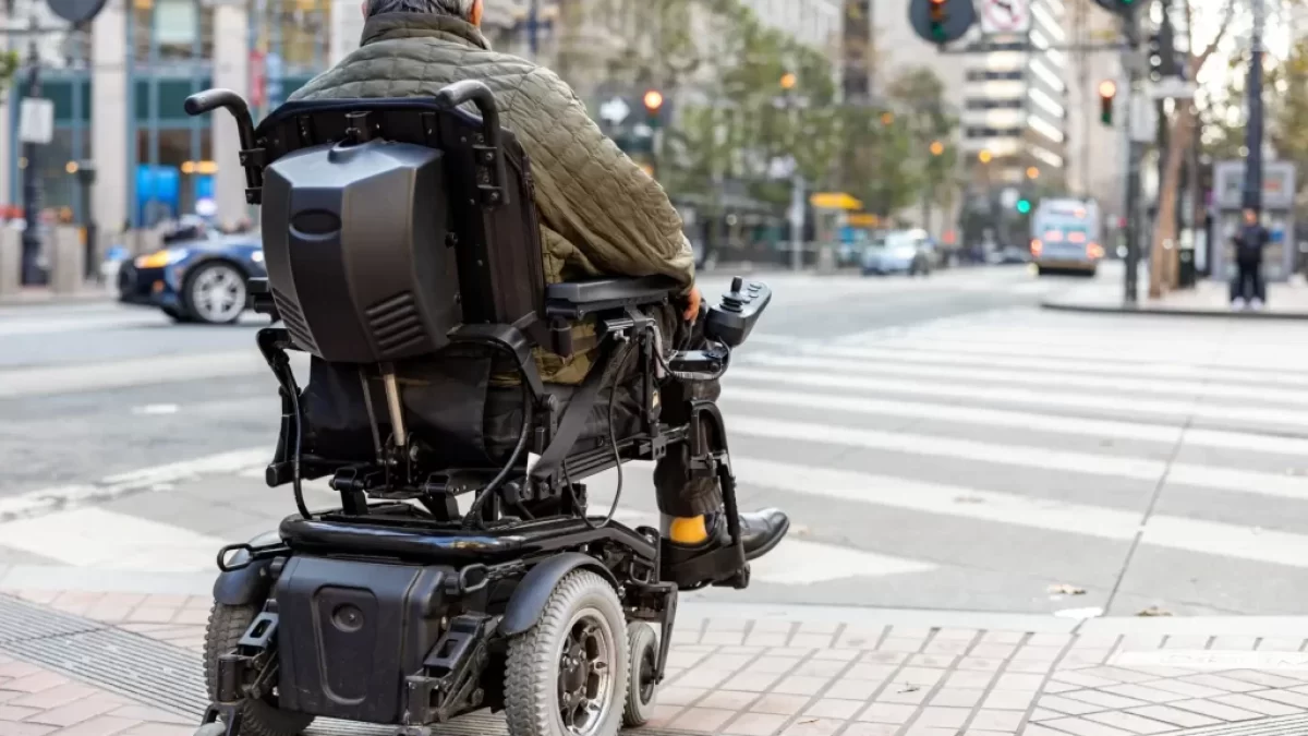 Starší muž sedící v elektrickém invalidním vozíku. Stojí na chodníku u přechodu pro chodce ve městě a čeká, až bude moci bezpečně přejet.