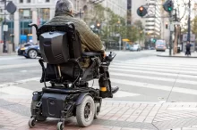 Starší muž sedící v elektrickém invalidním vozíku. Stojí na chodníku u přechodu pro chodce ve městě a čeká, až bude moci bezpečně přejet.