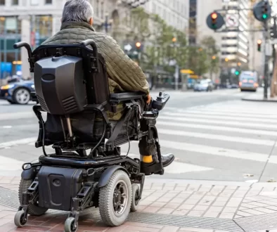 Starší muž sedící v elektrickém invalidním vozíku. Stojí na chodníku u přechodu pro chodce ve městě a čeká, až bude moci bezpečně přejet.