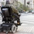 Starší muž sedící v elektrickém invalidním vozíku. Stojí na chodníku u přechodu pro chodce ve městě a čeká, až bude moci bezpečně přejet.