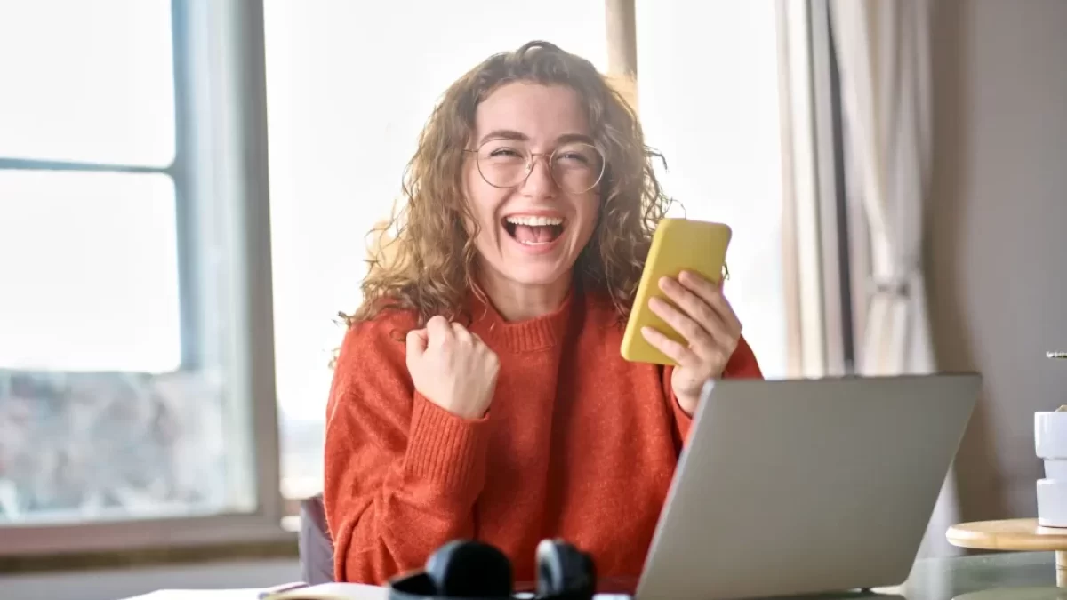 Mladá žena s kudrnatými vlasy, která sedí u stolu s notebookem a drží v ruce telefon.
