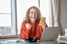 Mladá žena s kudrnatými vlasy, která sedí u stolu s notebookem a drží v ruce telefon.