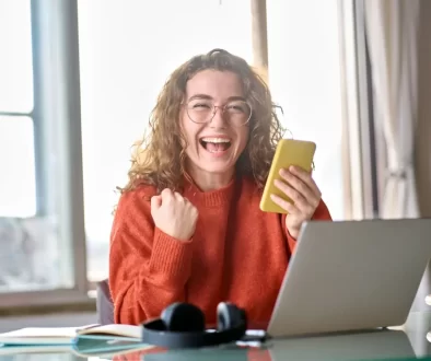 Mladá žena s kudrnatými vlasy, která sedí u stolu s notebookem a drží v ruce telefon.