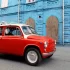 Veterán ZAZ-965 Záporožec v červeno-bílé barvě, zaparkovaný na ulici před výraznou modrou cihlovou budovou se třemi oblými dřevěnými vraty.