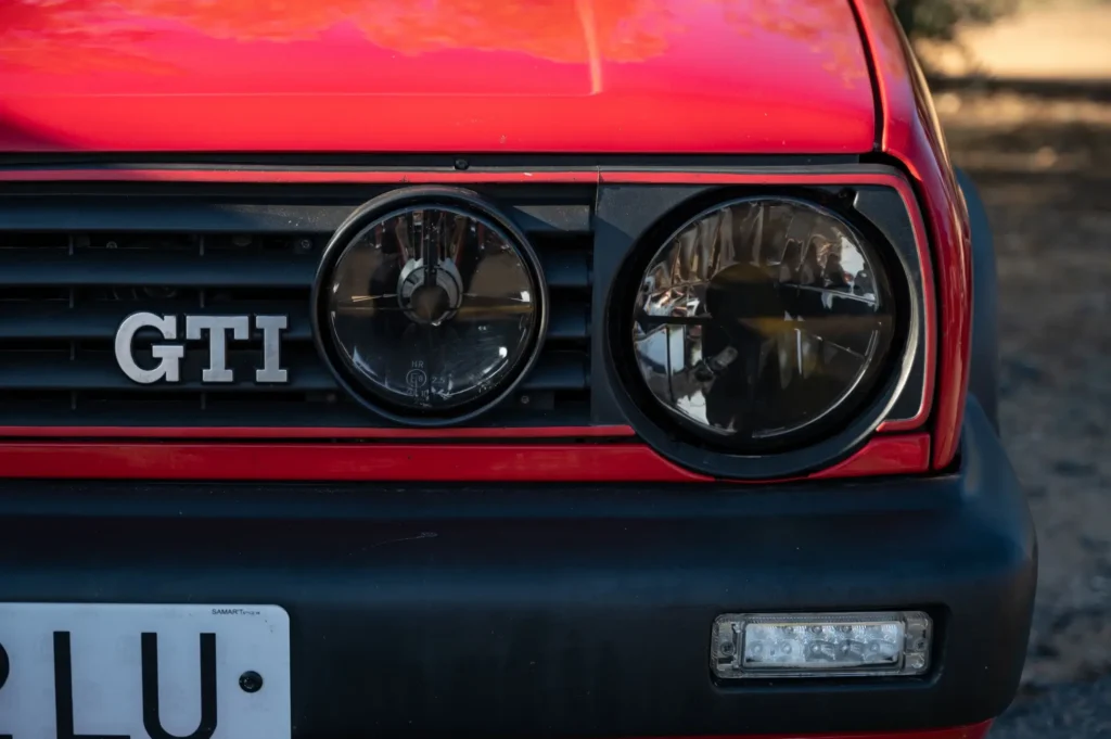 auto2 1 - Techsvět Detailní pohled na přední část Volkswagenu Golf GTI Mk2, konkrétně na dvojici kulatých světlometů, typickou červenou linku v masce chladiče a znak GTI.