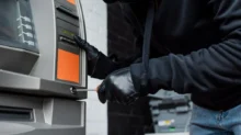 Člověk v černé kukle a rukavicích se pomocí šroubováku snaží upravit bankomat.