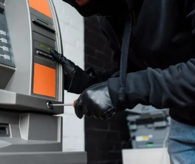Člověk v černé kukle a rukavicích se pomocí šroubováku snaží upravit bankomat.