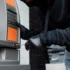 Člověk v černé kukle a rukavicích se pomocí šroubováku snaží upravit bankomat.