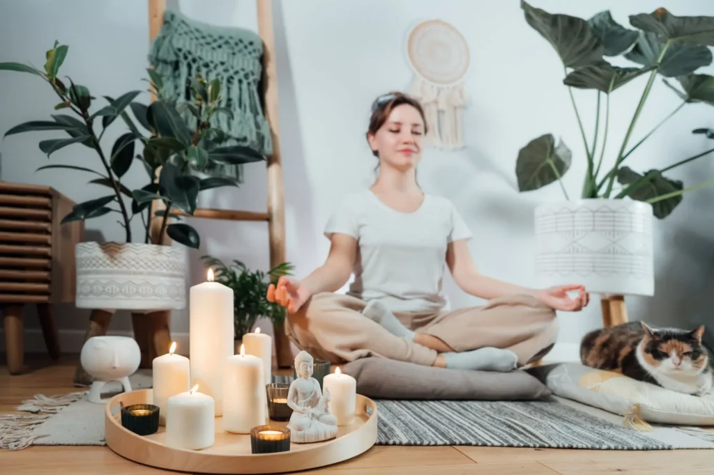 Žena v meditační pozici na podložce v harmonickém, útulném interiéru s pokojovými rostlinami, hořícími svíčkami, soškou Buddhy.