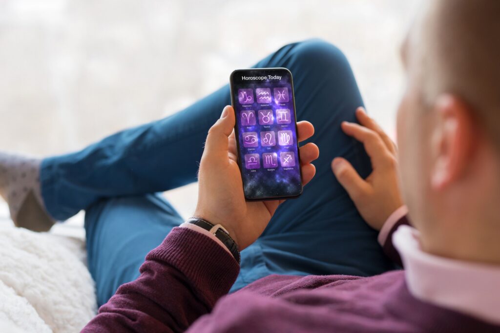 horoskop3 - Techsvět Muž pohodlně usazený na gauči, který drží v ruce mobilní telefon. Na displeji běží aplikace s názvem „Horoscope Today“, kde jsou zobrazeny všechny znamení zvěrokruhu ve fialových ikonách.