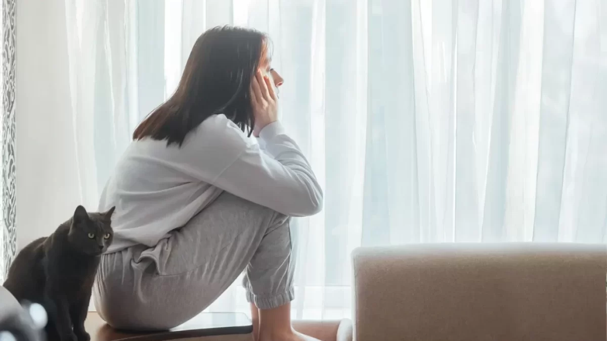 Žena sedící shrbená na parapetu u okna, zamyšleně hledí ven. Vedle ní sedí černá kočka, která se dívá stejným směrem.