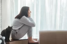 Žena sedící shrbená na parapetu u okna, zamyšleně hledí ven. Vedle ní sedí černá kočka, která se dívá stejným směrem.
