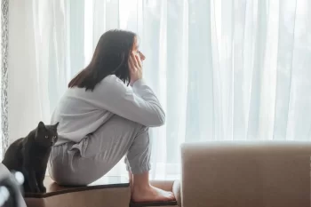 Žena sedící shrbená na parapetu u okna, zamyšleně hledí ven. Vedle ní sedí černá kočka, která se dívá stejným směrem.
