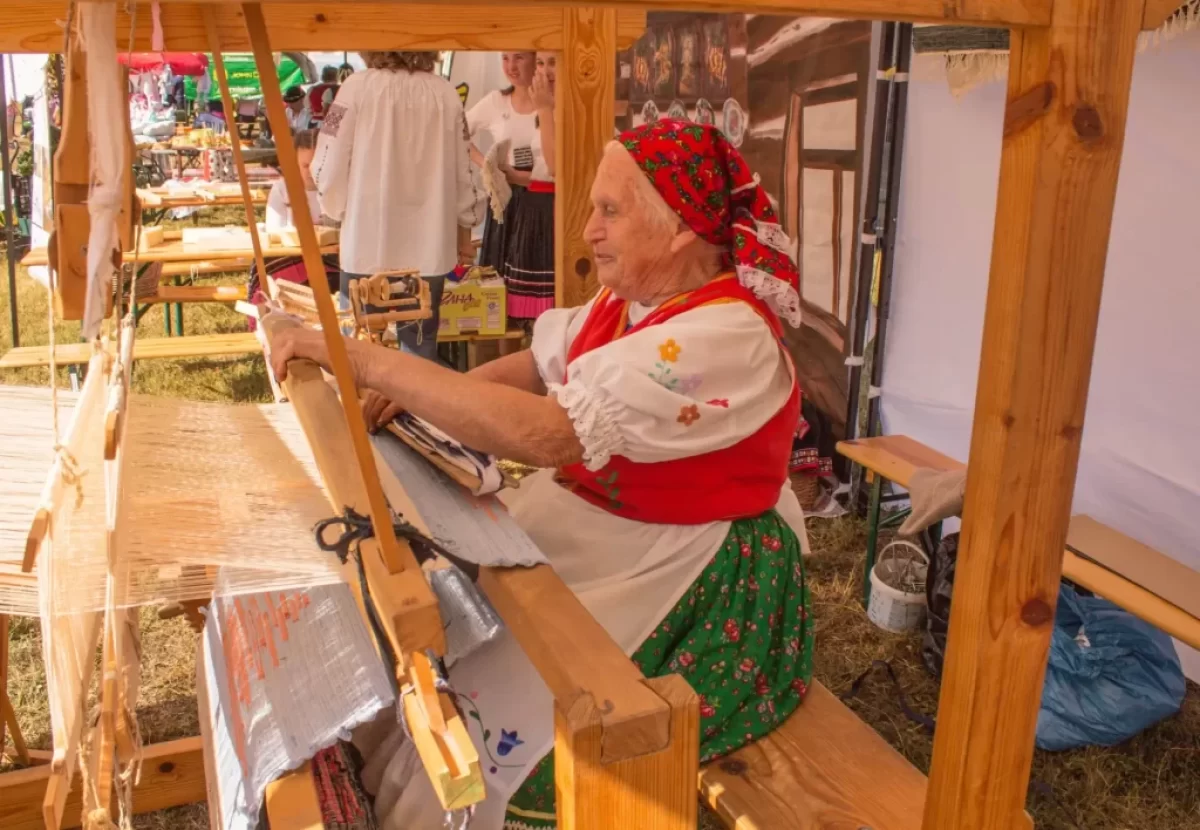 Starší žena v tradičním kroji, která tká na dřevěném stavu.