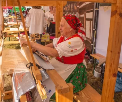 Starší žena v tradičním kroji, která tká na dřevěném stavu.