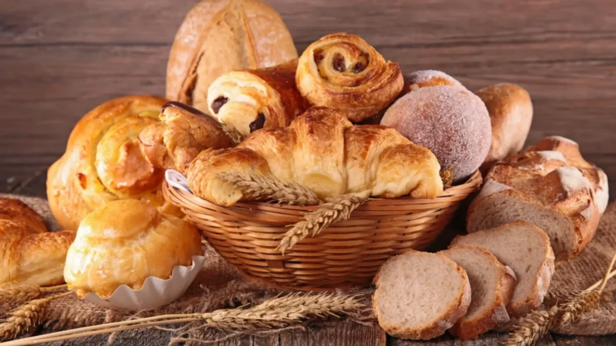 Proutěný koš plný různých druhů sladkého a slaného pečiva. Vidíme zde croissanty, šneky s rozinkami, koblihu obalenou cukrem, plněné koláče, máslové houstičky a několik krajíců čerstvě nakrájeného chleba.