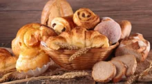 Proutěný koš plný různých druhů sladkého a slaného pečiva. Vidíme zde croissanty, šneky s rozinkami, koblihu obalenou cukrem, plněné koláče, máslové houstičky a několik krajíců čerstvě nakrájeného chleba.