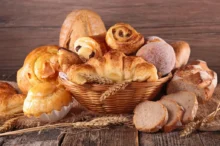 Proutěný koš plný různých druhů sladkého a slaného pečiva. Vidíme zde croissanty, šneky s rozinkami, koblihu obalenou cukrem, plněné koláče, máslové houstičky a několik krajíců čerstvě nakrájeného chleba.