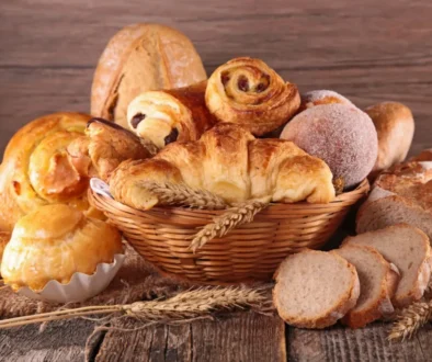 Proutěný koš plný různých druhů sladkého a slaného pečiva. Vidíme zde croissanty, šneky s rozinkami, koblihu obalenou cukrem, plněné koláče, máslové houstičky a několik krajíců čerstvě nakrájeného chleba.