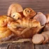 Proutěný koš plný různých druhů sladkého a slaného pečiva. Vidíme zde croissanty, šneky s rozinkami, koblihu obalenou cukrem, plněné koláče, máslové houstičky a několik krajíců čerstvě nakrájeného chleba.