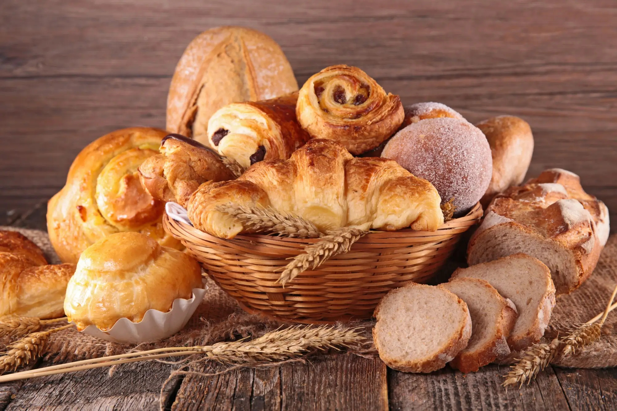 Proutěný koš plný různých druhů sladkého a slaného pečiva. Vidíme zde croissanty, šneky s rozinkami, koblihu obalenou cukrem, plněné koláče, máslové houstičky a několik krajíců čerstvě nakrájeného chleba.