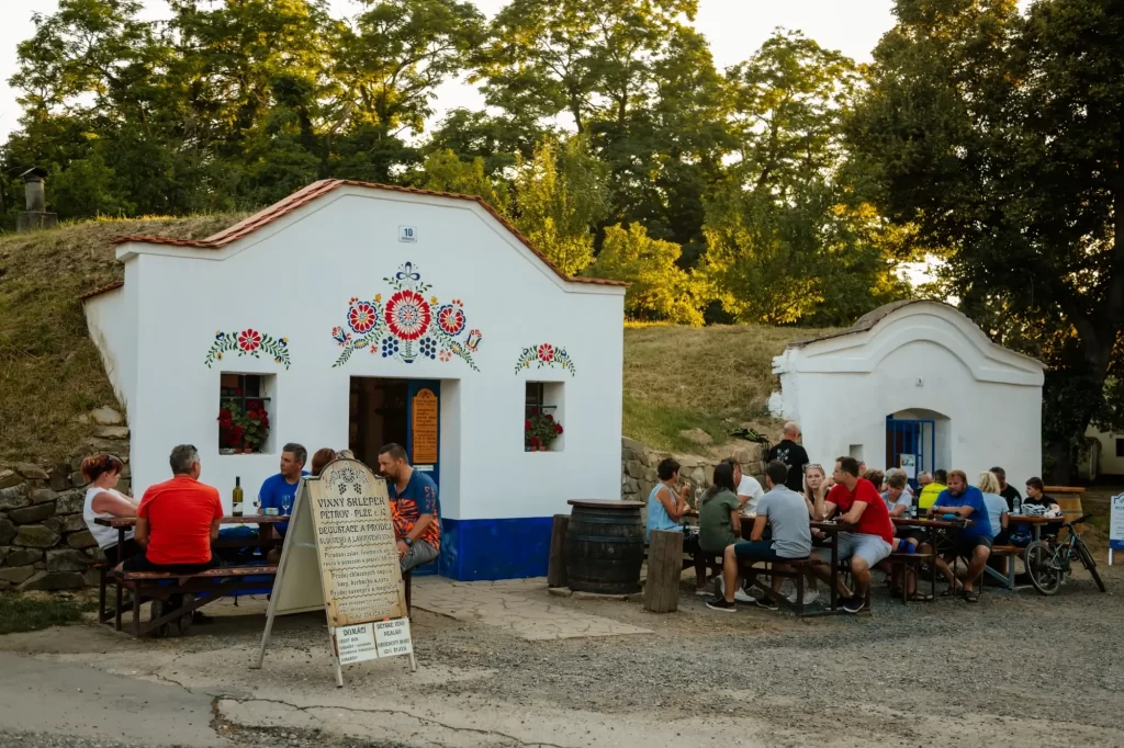 Posezení před tradičními vinnými sklepy v Plžích, kde skupiny lidí degustují víno a užívají si letní večer v malebném prostředí s folklórní výzdobou.