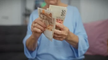 Starší žena v modré halence, která si doma prohlíží nebo přepočítává české bankovky.