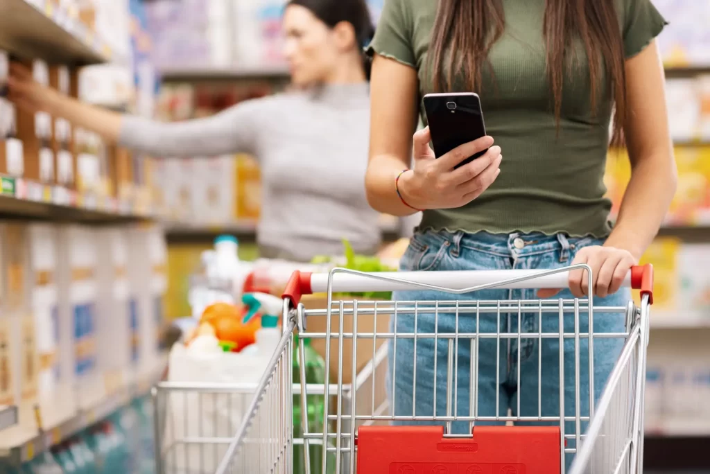 Mladá žena s nákupním vozíkem, která při nakupování v supermarketu používá mobilní telefon k porovnání cen.