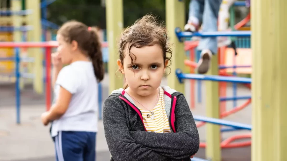 Malá holčička se zkříženýma rukama a vážným až naštvaným výrazem ve tváři. Stojí na dětském hřišti, kde si v pozadí hrají další děti.
