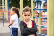 Malá holčička se zkříženýma rukama a vážným až naštvaným výrazem ve tváři. Stojí na dětském hřišti, kde si v pozadí hrají další děti.