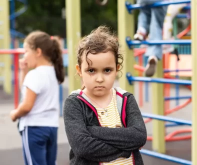 Malá holčička se zkříženýma rukama a vážným až naštvaným výrazem ve tváři. Stojí na dětském hřišti, kde si v pozadí hrají další děti.