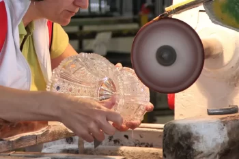 Detailní pohled na sklářku, která ručně brousí dekorativní křišťálovou mísu pomocí rotačního brusného kotouče.