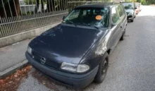 Modré auto stojí zaparkované na ulici, na čelním skle červená samolepka