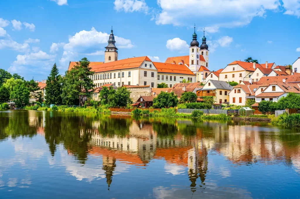 Panoramatický pohled na historické centrum Telče s dominantním zámkem a věžemi kostela, které se zrcadlí na hladině rybníka. 