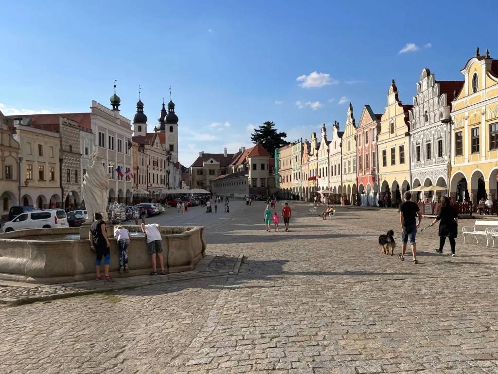 Hlavní náměstí v Telči s typickou řadou pestrobarevných historických domů, arkádami, kašnou v popředí a několika lidmi, kteří si užívají poklidné odpoledne. 