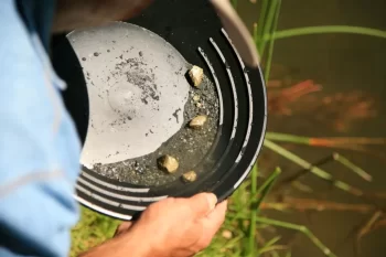 Detailní pohled na člověka, který drží rýžovací pánev nad vodou. V pánvi je vidět směs štěrku, černého písku a několik větších zlatavých kousků.