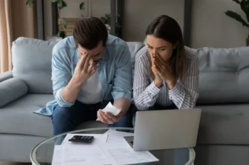 Nešťastný pár sedí v obývacím pokoji, obklopený rozházenými bankovními výpisy a smlouvami, s frustrovanými výrazy v obličeji