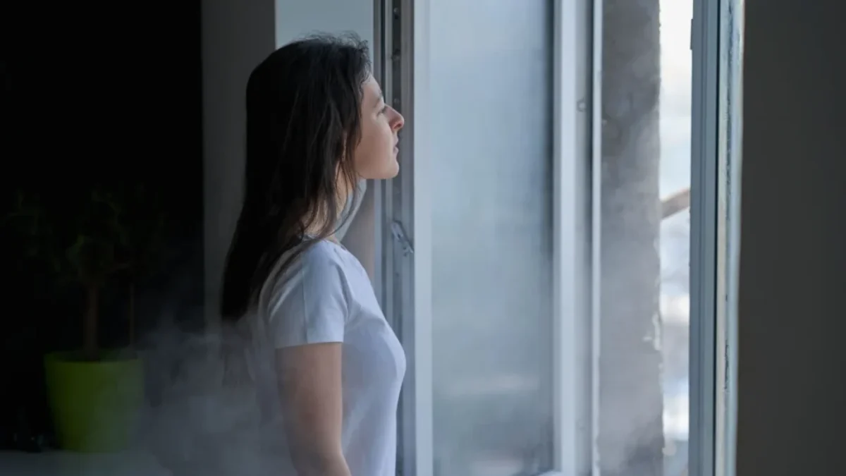 Mladá žena s hnědými vlasy a v bílém tričku stojí u otevřeného okna. Je zima, protože z venku do místnosti očividně proudí studený vzduch.