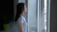 Mladá žena s hnědými vlasy a v bílém tričku stojí u otevřeného okna. Je zima, protože z venku do místnosti očividně proudí studený vzduch.