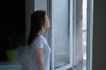 Mladá žena s hnědými vlasy a v bílém tričku stojí u otevřeného okna. Je zima, protože z venku do místnosti očividně proudí studený vzduch.