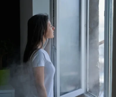 Mladá žena s hnědými vlasy a v bílém tričku stojí u otevřeného okna. Je zima, protože z venku do místnosti očividně proudí studený vzduch.