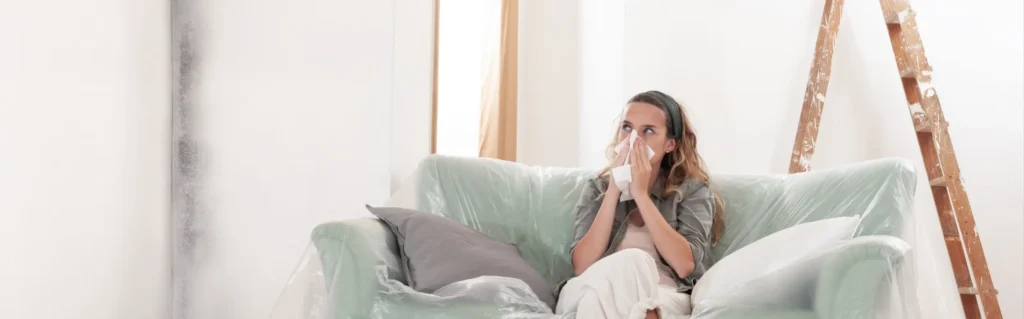 Mladá žena sedí na šedém gauči. U nosu si drží kapesník, má alergii na plíseň. Ta jde vidět v levém rohu místnosti.