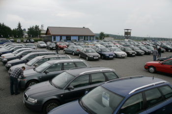 Large parking lot filled with rows of cars, a blue-roofed building in the background, and a wooden observation tower on the right, with a few people inspecting vehicles.