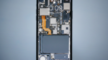 Close-up of a smartphone with its back removed, exposing motherboard, circuits, and orange flex cable along the side.