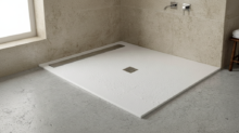 White rectangular shower base with a square drain in a minimalist bathroom, beige tiled walls, and wall-mounted faucets to the right.