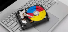 Hard disk drive resting on a silver laptop keyboard with a multicolored, Google-style circular label on the platter.
