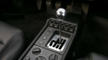 Interior of a black car showing a chrome manual gear shifter, center console with buttons and climate controls.
